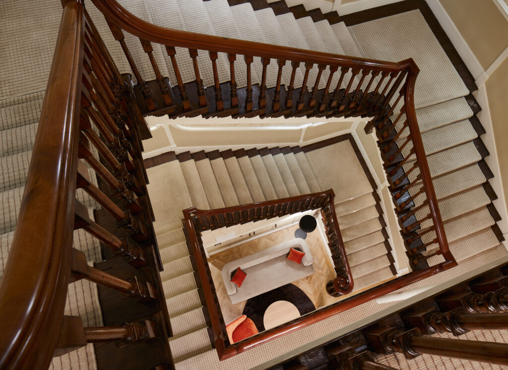 View from above of a grand wooden staircase spiraling downwards, highlighting classic architectural detailing and a cozy lounge area below.