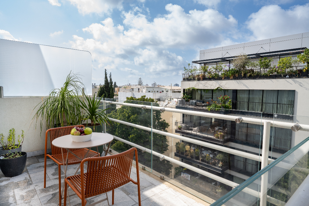 Sunny private balcony at master Mazeh featuring two orange chairs, a small table with a bowl of fruit, potted greenery, and views of surrounding modern residential buildings in Tel Aviv.