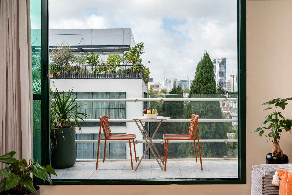 View from the apartment’s interior looking out onto a private balcony with two chairs, a fruit bowl on the table, green plants, and city skyline framed by tall trees.