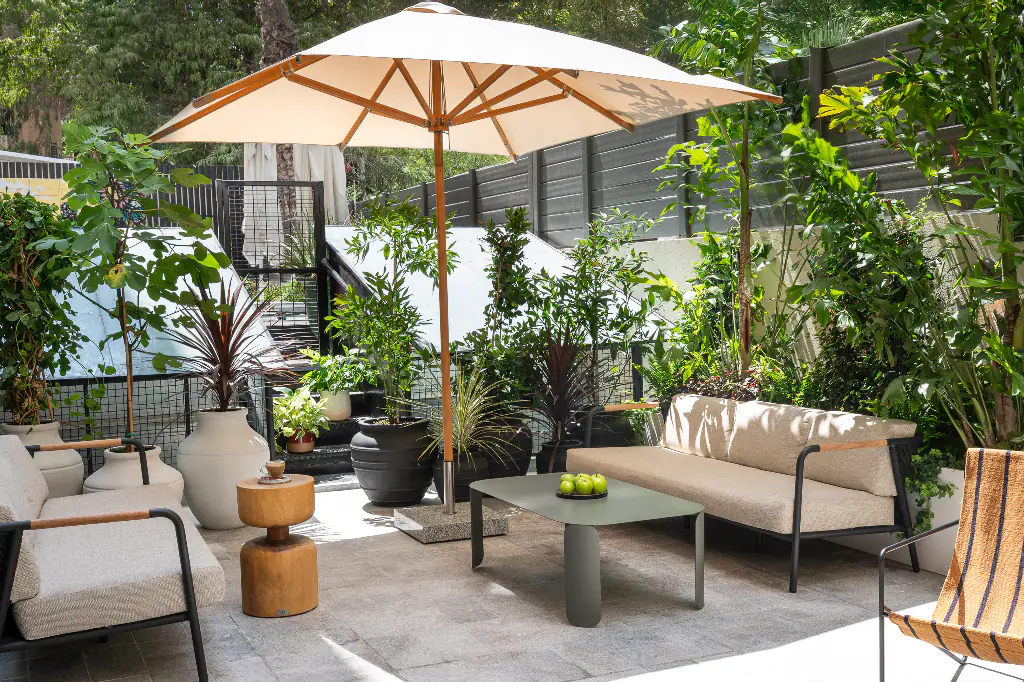 Lush garden terrace at master Mazeh with modern cushioned seating, potted plants, a central coffee table with green apples, and a large umbrella providing shade in a peaceful outdoor setting.