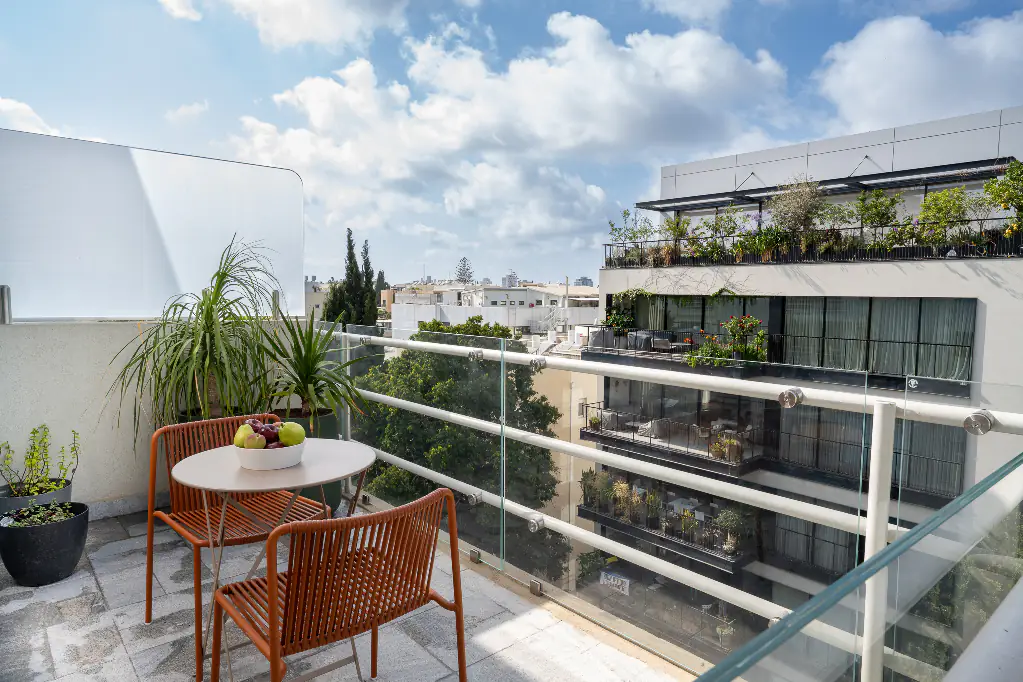 Sunny private balcony at master Mazeh featuring two orange chairs, a small table with a bowl of fruit, potted greenery, and views of surrounding modern residential buildings in Tel Aviv.