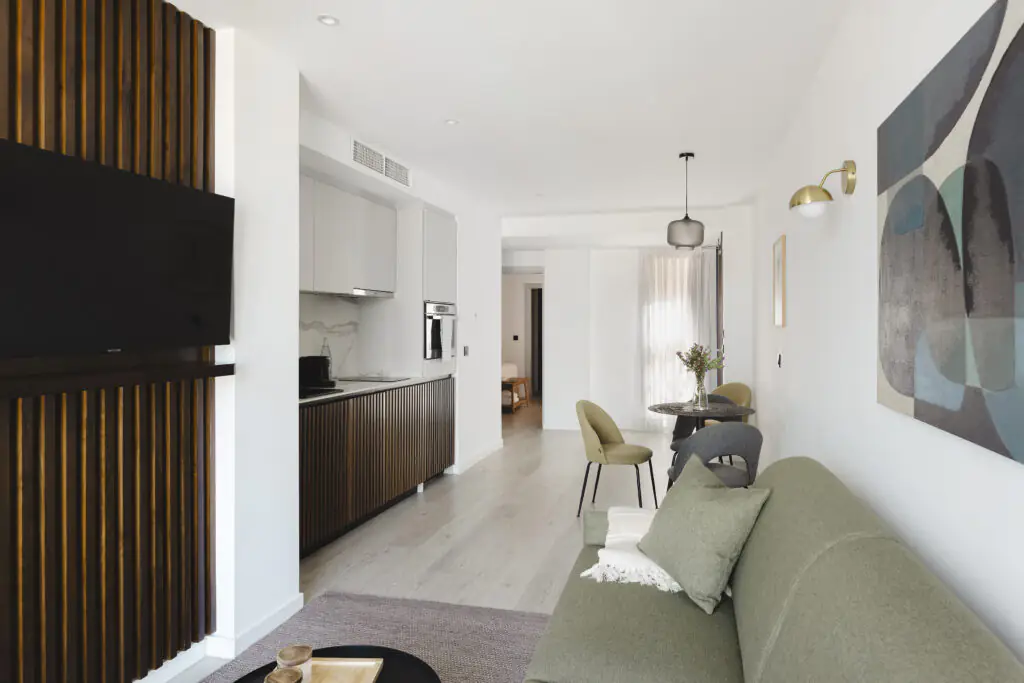 Modern open-plan living area at master La Rambla Barcelona with green sofa, flat-screen TV, and sleek kitchen design.